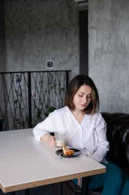 Kafede beyaz gömlekli, kendine güvenen esmer bir kadının portresi. Yanında bir fincan kahve ve tatlı. Bir dilim pasta..