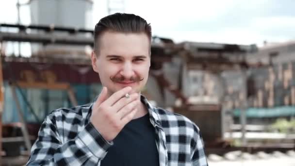 Jovem homem bonito com bigode em xadrez camisa preta e branca fumando um cigarro fora. mau hábito — Vídeo de Stock