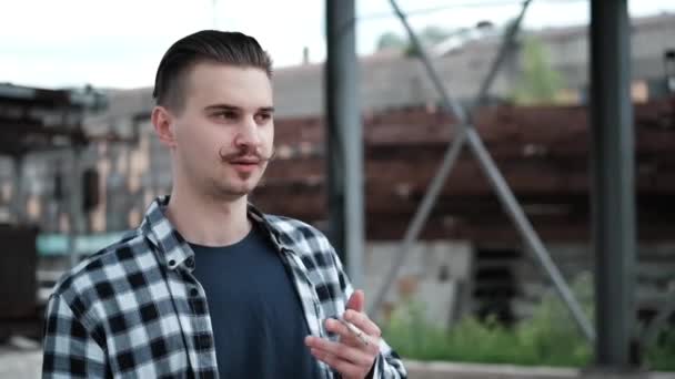 Jovem homem bonito com bigode em xadrez camisa preta e branca fumando um cigarro fora. mau hábito — Vídeo de Stock