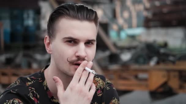 Молодой красивый мужчина с усами в черно-белой клетчатой рубашке курит сигарету на улице. вредная привычка — стоковое видео
