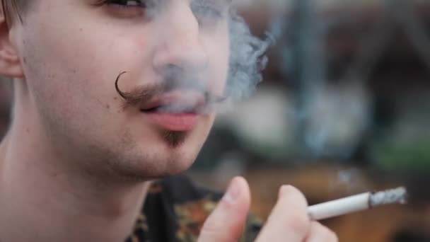 Jovem homem bonito com bigode em xadrez camisa preta e branca fumando um cigarro fora. mau hábito — Vídeo de Stock
