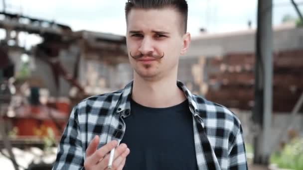 Jonge knappe man met snor in geruit zwart-wit shirt die buiten een sigaret rookt. slechte gewoonte — Stockvideo