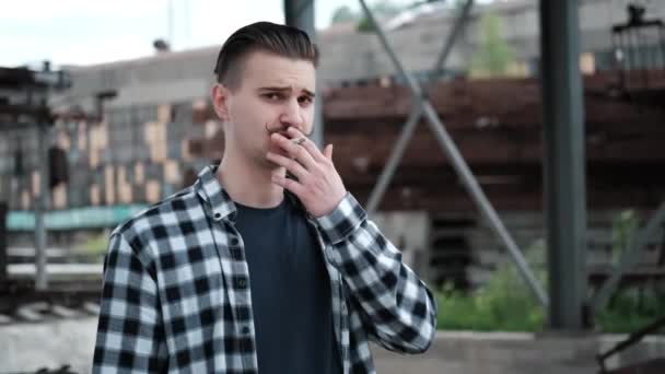 Joven hombre guapo con bigote en camisa a cuadros blanco y negro fumando un cigarrillo fuera. mal hábito — Vídeo de stock