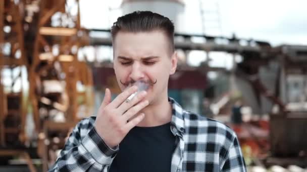 Jovem homem bonito com bigode em xadrez camisa preta e branca fumando um cigarro fora. mau hábito — Vídeo de Stock