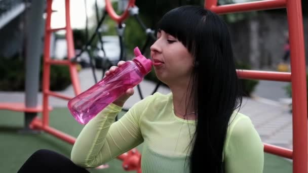 Moda, siyah ve yeşil spor kıyafetleri içindeki çekici fitness kızı. Spor sahasında pembe bir şişe su ile sokakta moda olan çocuk parkında. — Stok video