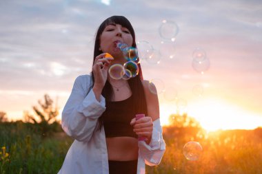 Gün batımında yeşil yaz tarlasında sabun köpüğü yapan güzel genç bir kadın. Mutlu barışçıl zamanlar. Milenyum nesli.