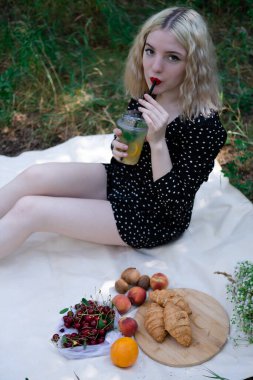 Ormanda piknik yapan güzel sarışın kız ya da limonata, meyve ve kruvasanla parkta. Yaz, dinlenme, rahatlama. Milenyum nesli.