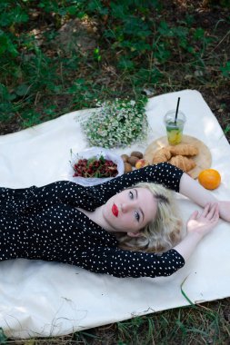 Parkta beyaz ekose üzerinde piknik yapan güzel sarışın bir kız. Elinde limonatalı şeffaf plastik bir bardak var. Taze yaz ferahlatıcı içeceği.