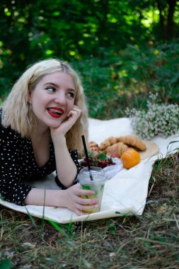 Parkta ekose bir piknikte lezzetli atıştırmalıklarla güzel sarışın bir kadın. Limonata, meyve ve kruvasan. Yaz zamanı, dinlen, rahatla, tadını çıkar. özgürlük.
