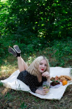 Ormanda piknik yapan güzel sarışın kız ya da limonata, meyve ve kruvasanla parkta. Yaz, dinlenme, rahatlama. Milenyum nesli.