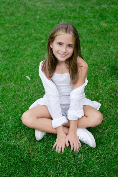 pretty tween girl in white clothes sitting on green grass outside in park on sunny summer day. charming ukrainian child portrait.