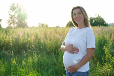 Güzel hamilelik portresi. Mutlu bir anne, gün batımında beyaz tişörtlü ve kot pantolonlu bir şekilde çayırda olduğu için mutlu..