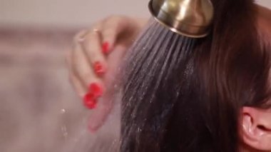 Genç esmer kadın uzun saçlarını duşun altında yıkıyor sırtı kameraya dönük duruyor ve su püskürtüsünün altında temizliyor. Modern bej banyoda..