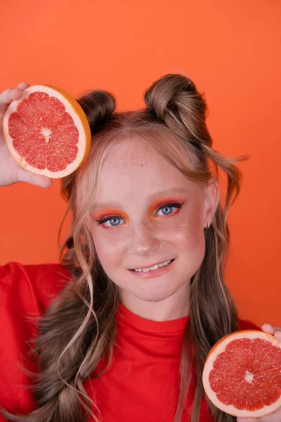 オレンジの背景にグレープフルーツのスライスを持つ若い女の子 巻き毛と2本のポニーテール — ストック写真