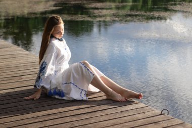 a girl in an embroidered Ukrainian shirt sits on the pier, the reflection of clouds in the water of the lake. On the shore of the sky. vyshyvanka day. freedom. patriot.