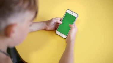 teenager holding smartphone in hands with green screen for keying. Over the shoulder view from back of child having video chat, playing, studying on chroma key. boy gadget digital phone. Slow motion.