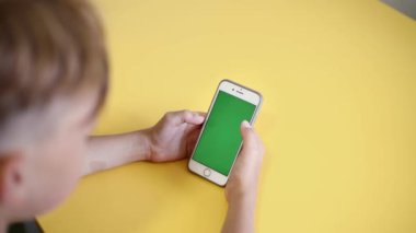 teenager holding smartphone in hands with green screen for keying. Over the shoulder view from back of child having video chat, playing, studying on chroma key. boy gadget digital phone. Slow motion.