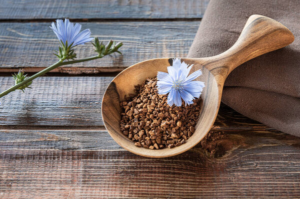 Close-up of instant chicory granules and fresh blue flower in wooden spoon on rough table made of brown natural wooden planks with cracks. Linen textile napkin and plant stem in bloom on side.