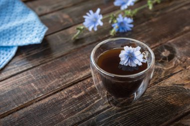 Şeffaf termo cam bardaktaki hindiba, çiçek açan hindiba kökleri ve mavi keten peçeteden yapılmış kahve içeceğinin alternatifi. Arka planda ahşap tahtalardan yapılmış sert kahverengi masa yüzeyi. Boşluğu kopyala.