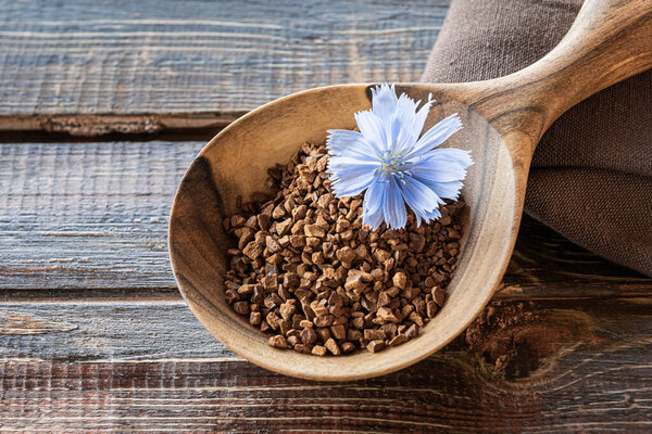 Close-up of instant chicory granules and fresh blue flower in large handcrafted spoon on rough table made of brown natural wooden planks with cracks. Linen textile napkin folded on side. Copy space.