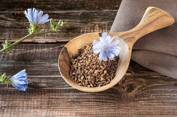 Fresh blue flower and Instant granules of chicory in wooden spoon on rough table made of brown natural wooden planks with cracks. Linen textile napkin and plant stems in bloom on side. Close-up.