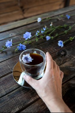 Elinde hindiba kökü ve termo cam bardaktan yapılmış hindiba kökü olan beyaz bir kadın eli ve kahverengi ahşap masa üzerinde çiçek açan hindiba kökleri. Kahveye alternatif kafein. Uzayı kopyala Kırsal biçim.