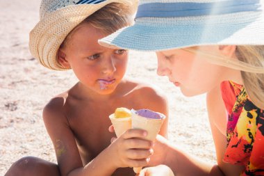 Havada dinlenen sarışın bir oğlan ve şapkalı sarışın bir kız kumsalda, kumsalda oturuyor ve dondurma yiyor ya da tadıyor küçük bir tarla derinliği ve kopyalama alanı ile. Tatil kavramı  