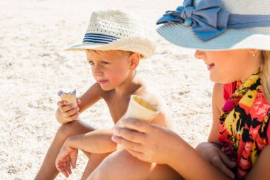 Havada dinlenen sarışın bir oğlan ve şapkalı sarışın bir kız kumsalda, kumsalda oturuyor ve dondurma yiyor ya da tadıyor küçük bir tarla derinliği ve kopyalama alanı ile. Tatil kavramı  