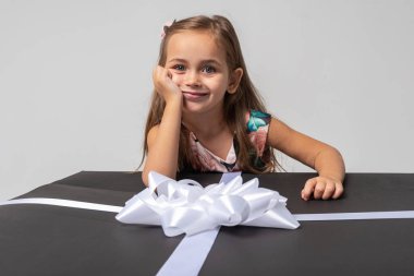 Siyah saçlı, renkli elbiseli, beyaz bir arka planda fiyonklu siyah bir kutuya yaslanmış küçük, tatlı, duygusal bir kız. Düşünceli ve gülümsüyor. Doğum günü, kutlama, beklenti.