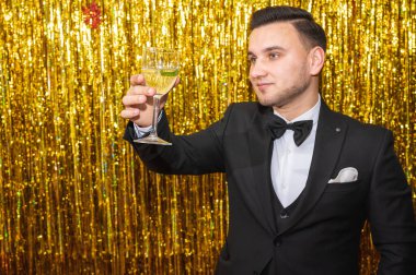 Siyah takım elbiseli şık bir genç adam altın duvarın yanında bir kelebek tutuyor ve bir bardak içkiyi kaldırıyor, neşeli bir ruh hali, gülümsüyor, başka tarafa bakıyor. Tatil, parti konsepti. Seçici odak