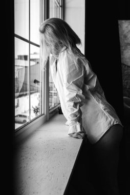 Portrait of a black and white young, pretty European girl emotional in a men's shirt in the bedroom near a large window, smiles, straightens hair. Morning sun, selective focus 