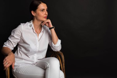 Portre, stüdyo çekimi, gülümseyen güzel, duygusal, çekici orta yaşlı bir kız karanlık bir arka planda bir sandalyede oturuyor.