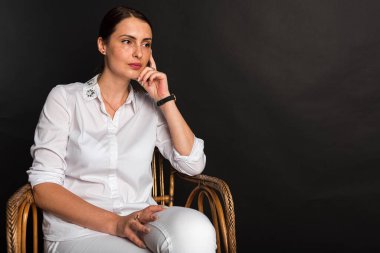 Portre, stüdyo çekimi, gülümseyen güzel, duygusal, çekici orta yaşlı bir kız karanlık bir arka planda bir sandalyede oturuyor.