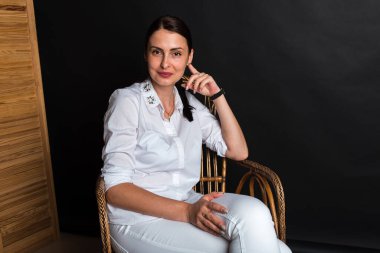 Portre, stüdyo çekimi, gülümseyen güzel, duygusal, çekici orta yaşlı bir kız karanlık bir arka planda bir sandalyede oturuyor.