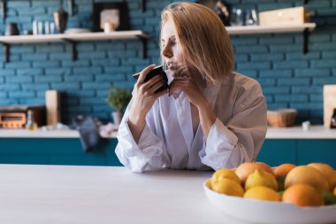Young beautiful European woman in a white men's shirt sitting with a cup of tea, coffee at the table in the kitchen at home. Morning coffee, tea 