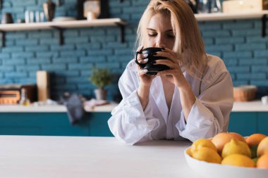 Young beautiful European woman in a white men's shirt sitting with a cup of tea, coffee at the table in the kitchen at home. Morning coffee, tea 