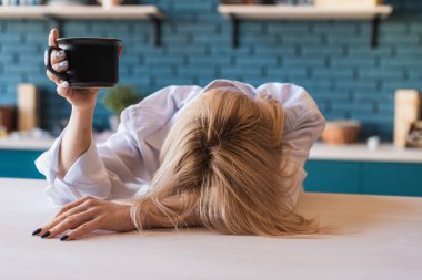 Yorgun genç sarışın kız mutfak masasına başını eğdi ama elinde koca bir fincan kahve tutuyordu.