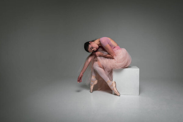 Young pretty, fragile, beautiful ballerina sits tired, tortured in a long pale pink dress with tulle on a uniform background, hand movements, restrained tone. Ballet, dance, dancer.