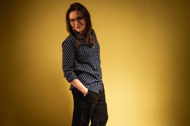 Koyu renk pantolonlu, gömlekli, duygusal, gülümseyen bir kadın. Örgü gömlekli çekici kız sarı arka planda dans ediyor. Genç bir kızın portresi. Yazı için yer