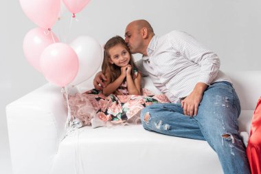 Babam, açık arkaplanda, beyaz bir kanepenin üzerinde oturan, sarı saçlı, küçük, güzel bir kızın yanında oturuyor. O gülümsüyor. Doğum günü, tatil