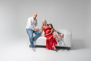 Kırmızı elbiseli güzel hamile bir anne ve beyaz arka planda izole edilmiş beyaz bir kanepede sevimli ve duygusal bir kız olan bir baba. Aile, bebek doğumu kavramı
