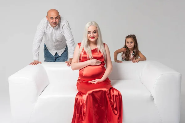 白い背景に隔離された白いソファの上に可愛くて感情的な女の子を持つ赤いドレスと父親の美しい妊婦の母親 出産の概念 — ストック写真