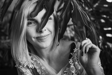 Genç, güzel bir kız, pencerenin yanında tropik çiçekleri, yeşil yaprakları, kanepeye yaslanmış, organik cilt bakımı, lüks spa oteli. Siyah beyaz klasik fotoğraf. Seçici odak