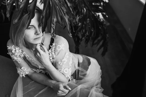 Genç, güzel bir kız, pencerenin yanında tropik çiçekleri, yeşil yaprakları, kanepeye yaslanmış, organik cilt bakımı, lüks spa oteli. Siyah beyaz klasik fotoğraf. Seçici odak