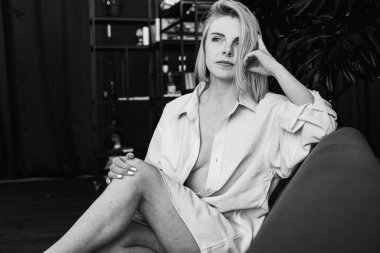 Siyah beyaz klasik fotoğraf, genç sarışın, pencerenin yanındaki sandalyede oturan güzel kız tropik çiçekli beyaz gömlek, organik cilt bakımı, lüks spa oteli, yaşam tarzı fotoğrafı. Seçici odak