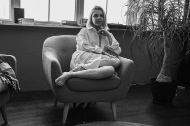 Siyah beyaz klasik fotoğraf, genç sarışın, pencerenin yanındaki sandalyede oturan güzel kız tropik çiçekli beyaz gömlek, organik cilt bakımı, lüks spa oteli, yaşam tarzı fotoğrafı. Seçici odak