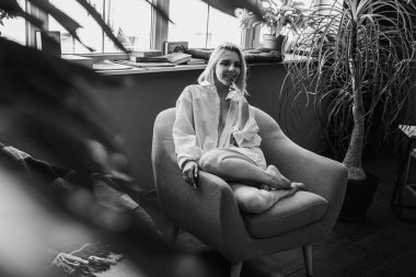 Siyah beyaz klasik fotoğraf, genç sarışın, pencerenin yanındaki sandalyede oturan güzel kız tropik çiçekli beyaz gömlek, organik cilt bakımı, lüks spa oteli, yaşam tarzı fotoğrafı. Seçici odak