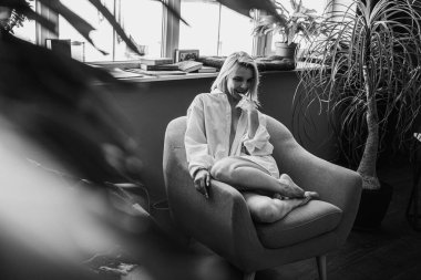 Siyah beyaz klasik fotoğraf, genç sarışın, pencerenin yanındaki sandalyede oturan güzel kız tropik çiçekli beyaz gömlek, organik cilt bakımı, lüks spa oteli, yaşam tarzı fotoğrafı. Seçici odak