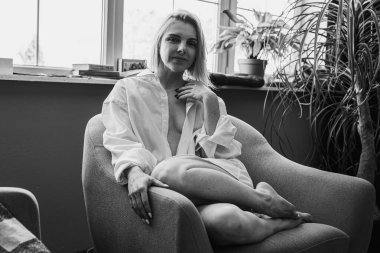 Siyah beyaz klasik fotoğraf, genç sarışın, pencerenin yanındaki sandalyede oturan güzel kız tropik çiçekli beyaz gömlek, organik cilt bakımı, lüks spa oteli, yaşam tarzı fotoğrafı. Seçici odak