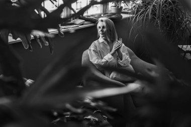 Siyah beyaz klasik fotoğraf, genç sarışın, pencerenin yanındaki sandalyede oturan güzel kız tropik çiçekli beyaz gömlek, organik cilt bakımı, lüks spa oteli, yaşam tarzı fotoğrafı. Seçici odak